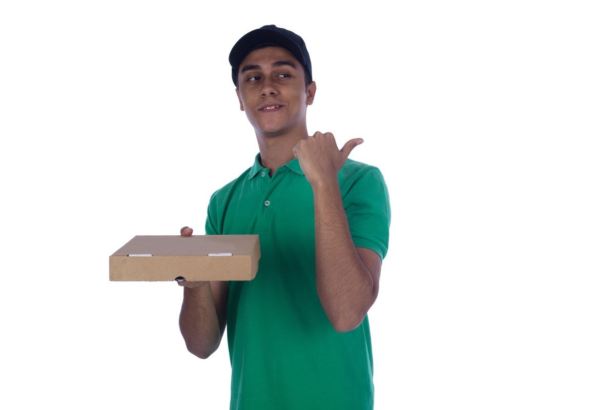 Young Delivery man holding closed pizza box pointing thumbs back with a smile wears green t shirt and cap isolated on white background.